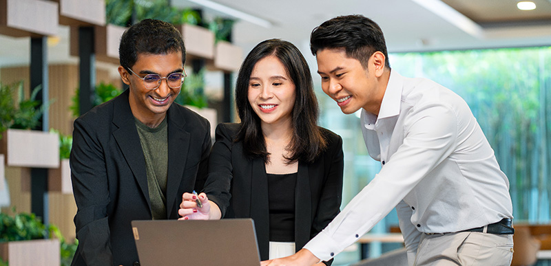 Three colleagues smiling while working together on a laptop in a modern office.